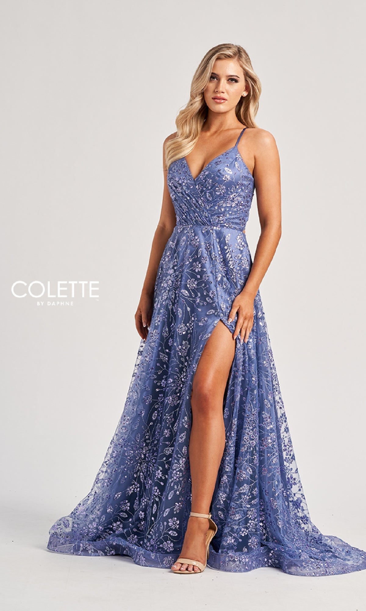 A woman wears the Colette CL8230 Long Prom Dress, a sleeveless, floor-length lavender gown with floral embellishments and a high slit, standing against a plain light background.